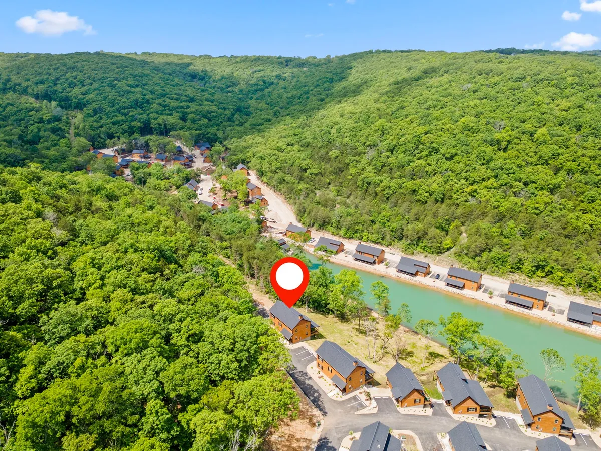 Wooden cabins sit near a river, surrounded by verdant hills and trees. The landscape is open and inviting, with the river hinting at fishing and boating opportunities. The scene is relaxed, evoking the slower pace of life and opportunity for early morning fishing or stargazing by the water.