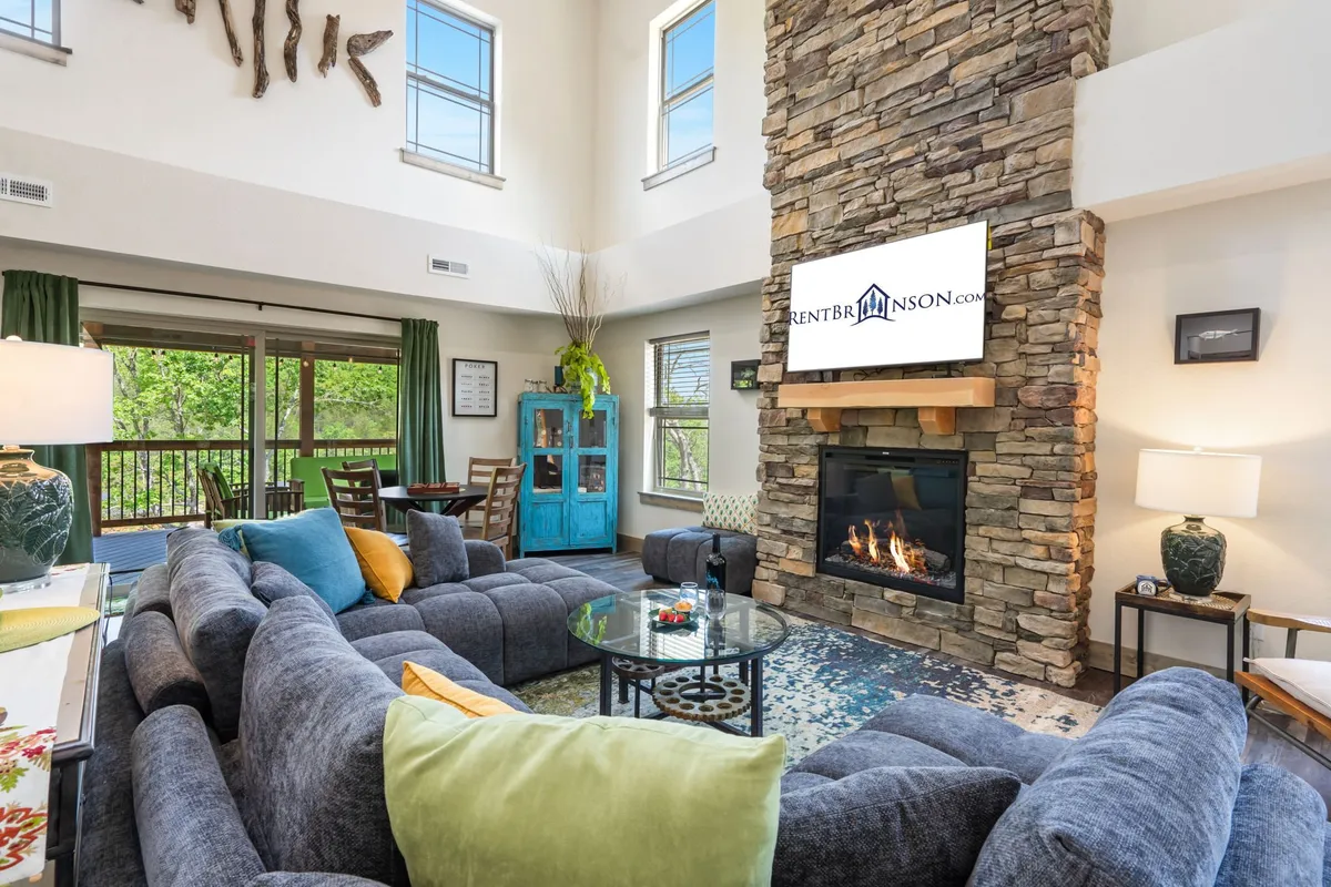 The image showcases a warm, open-concept living room featuring a striking stone fireplace as the centerpiece, an oversized sectional sofa perfect for gathering, a stylish blue cabinet, and a flat-screen TV, all set in a space designed for comfort and connection.