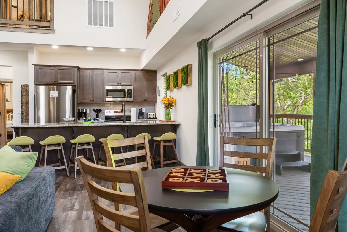 An inviting kitchen and dining space with a kitchen island surrounded by bar stools, a spacious dining table ready for meals or games, and large windows leading to an outdoor area. Decorative wall art and visible board games on the dining table add a playful, family-friendly touch.