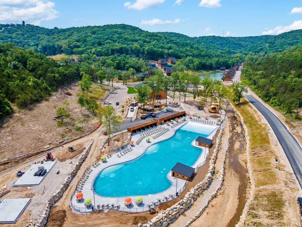 A large outdoor swimming pool sits amidst lush greenery, with buildings in the background and comfortable poolside amenities. The setting is ideal for families, with ample space for swimming, sunbathing, and social gatherings, capturing the energy and joy of summer days at the resort.