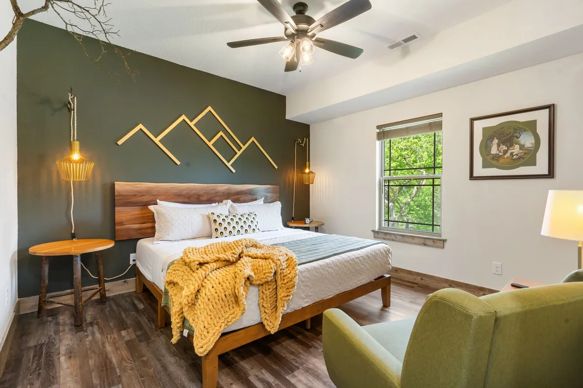 A tranquil bedroom featuring a bed with plush, decorative pillows, mountain-themed wall art, a green accent chair, wooden side table, and a decorative throw blanket. Sunlight filters softly through the window, creating a serene, inviting atmosphere.