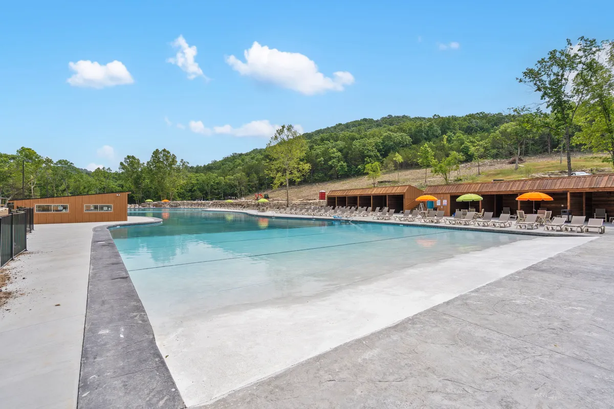 A large outdoor swimming pool surrounded by green hills, colorful umbrellas, and deck chairs. The pool area buzzes with activity, offering a scenic and leisurely spot for swimming, sunbathing, and gathering with friends and family.