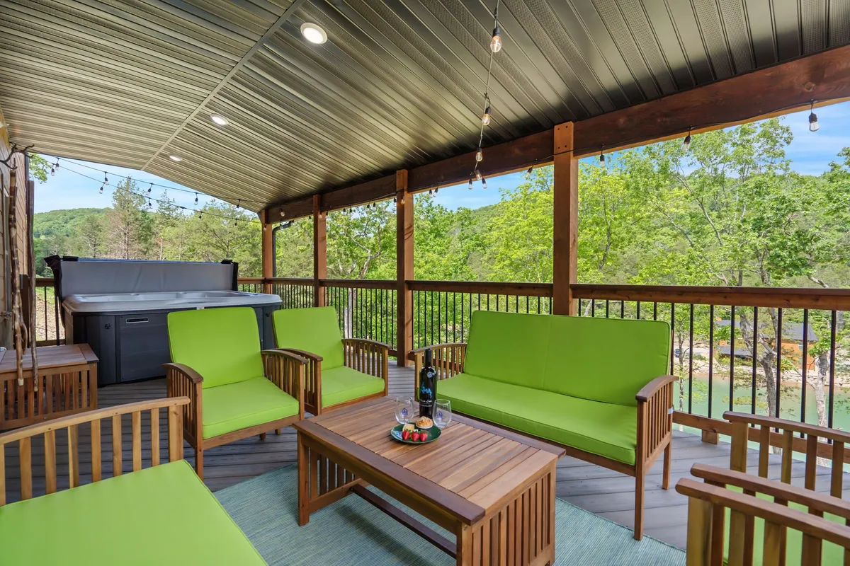 An exterior deck scene with plush green seating, a wooden table, a hot tub, and a natural view beyond the railings. The setup is perfect for gatherings, enjoying a drink, or quiet moments while stargazing or watching sunsets.