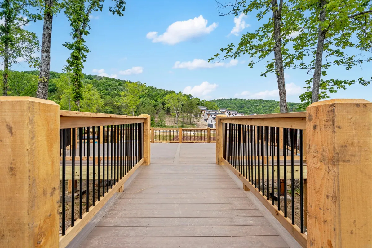 A wooden bridge stretches across a leafy area, framed by towering trees and hills in the distance. The setting is peaceful and untouched, inviting visitors to walk, photograph, and enjoy tranquil moments surrounded by nature. The pathway alludes to scenic trails awaiting exploration.